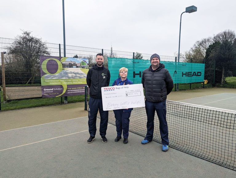 The photo shows coaches Ben Walker and Rob Hodge being presented with a cheque by Jacqueline from Tesco Stronger Starts on court at Wigginton Tennis Club